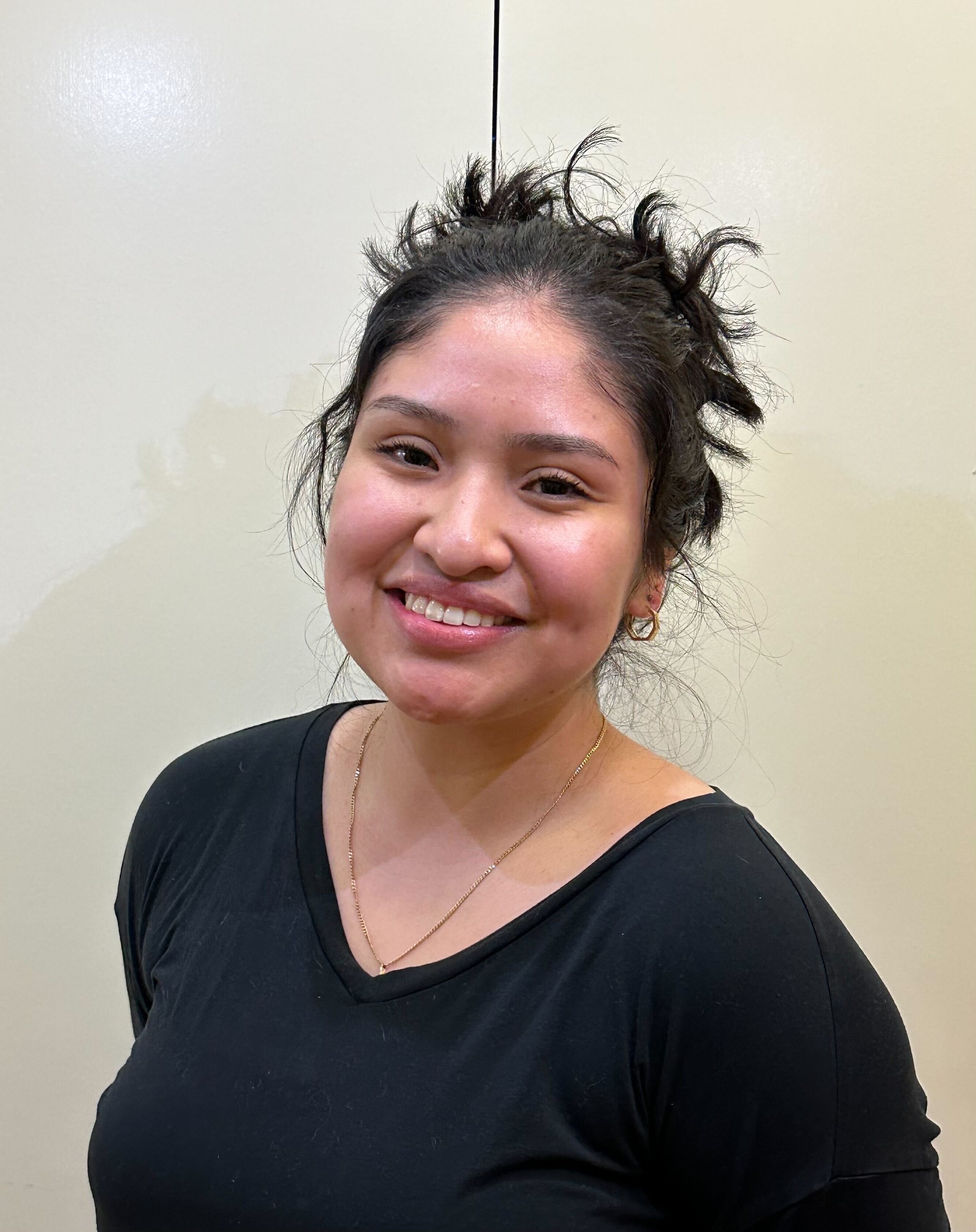 Paola Zavala is standing in the middle of the camera frame, facing the camera and smiling. 