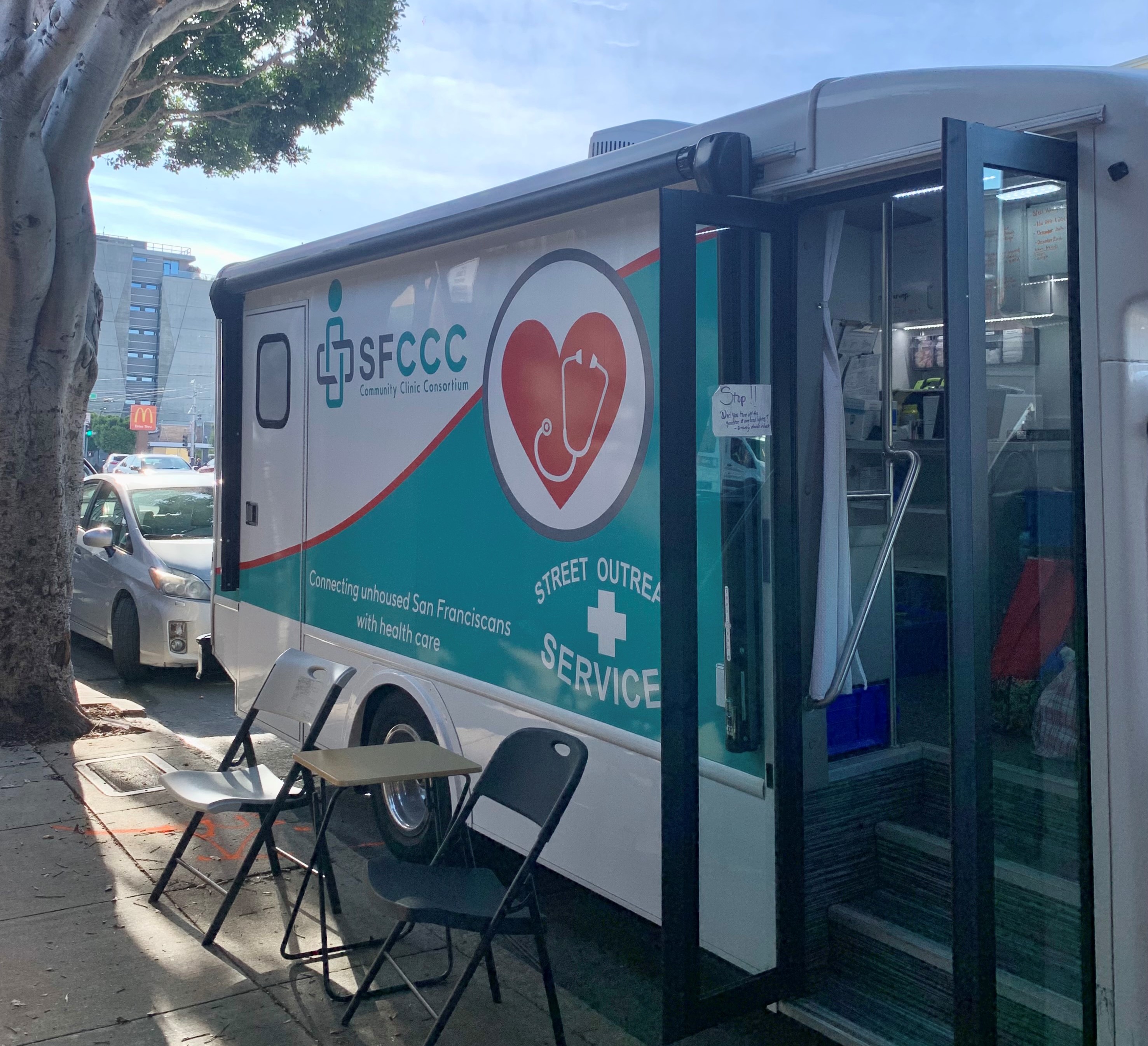 A photo of the Street Outreach Services (SOS) van, parked. There's a table with two folding chairs on either side of it. On the van, the sign reads "Connecting unhoused San Franciscans with health care."