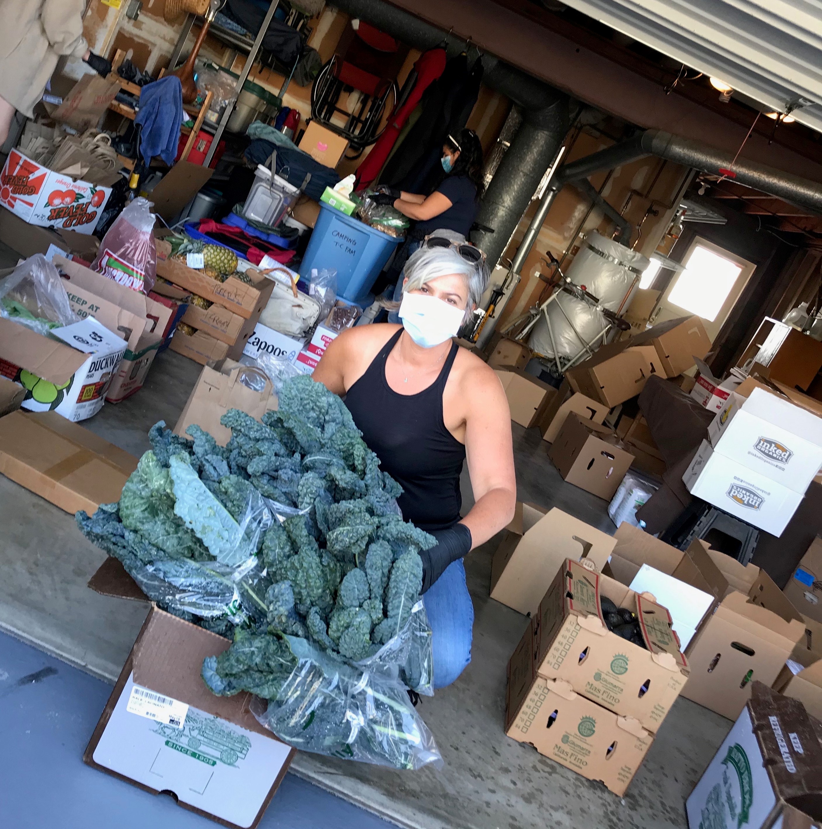 Janna is seen in the center of the frame, facing the camera with a mask on. She is squatting behind a box that contains kale, a green leafy vegetable to hand out to patients.
