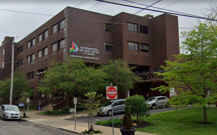 A brown brick building that says "The Behavioral Wellness Center at Girard" with green trees and some parked cars in the foreground