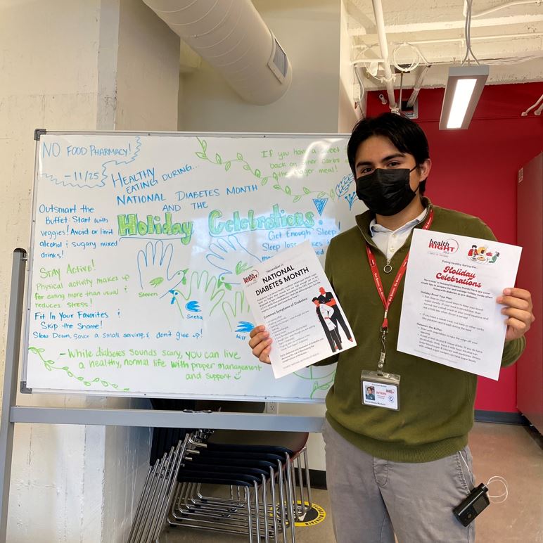Steven standing beside a white board about healthy eating during the holiday celebrations. Steven is holding up educational material for his Host Site, HealthRIGHT 360, about managing diabetes during the holidays.