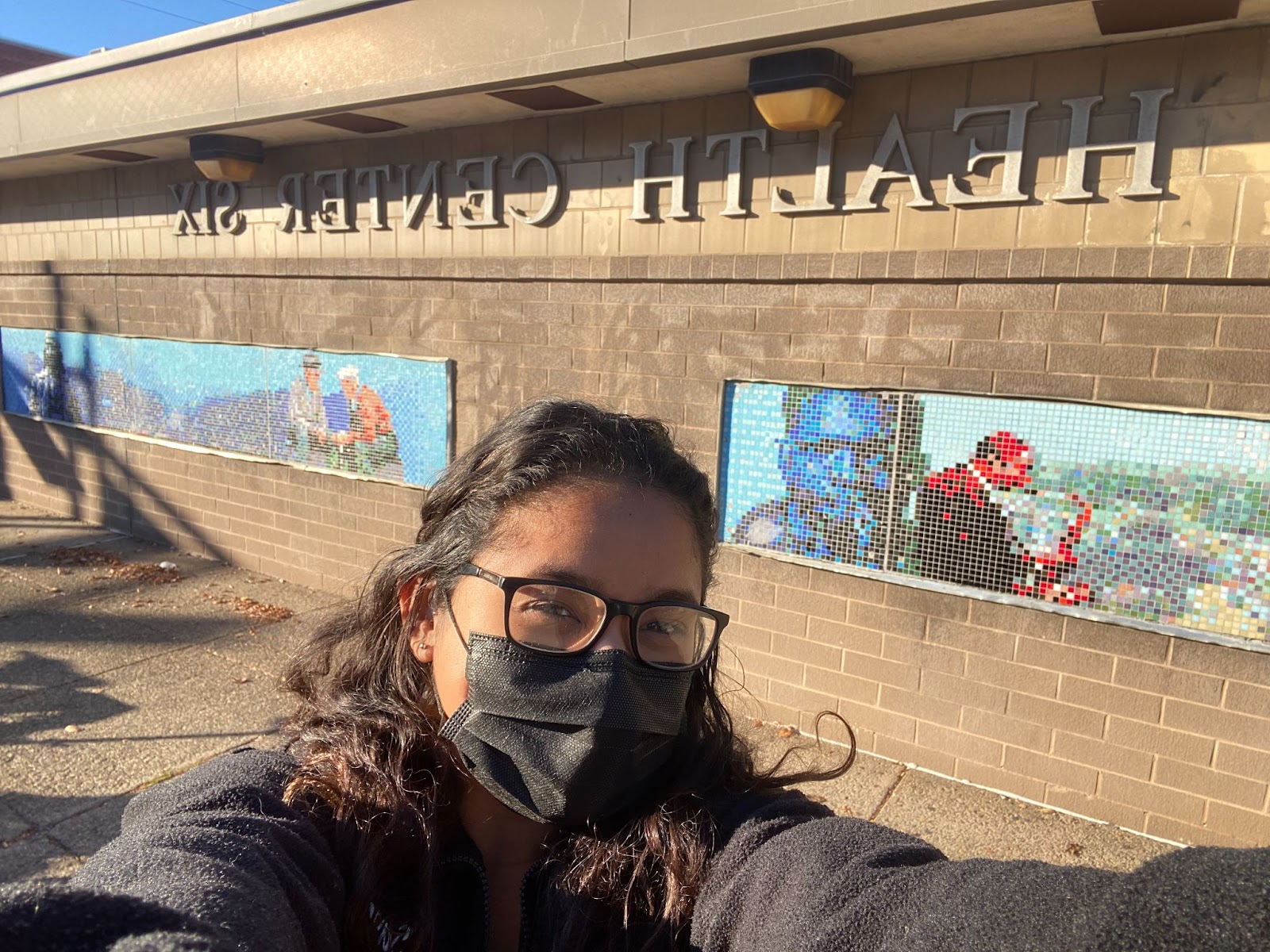 A selfie of Aliza wearing a mask and glasses standing outside of a building that says health center six