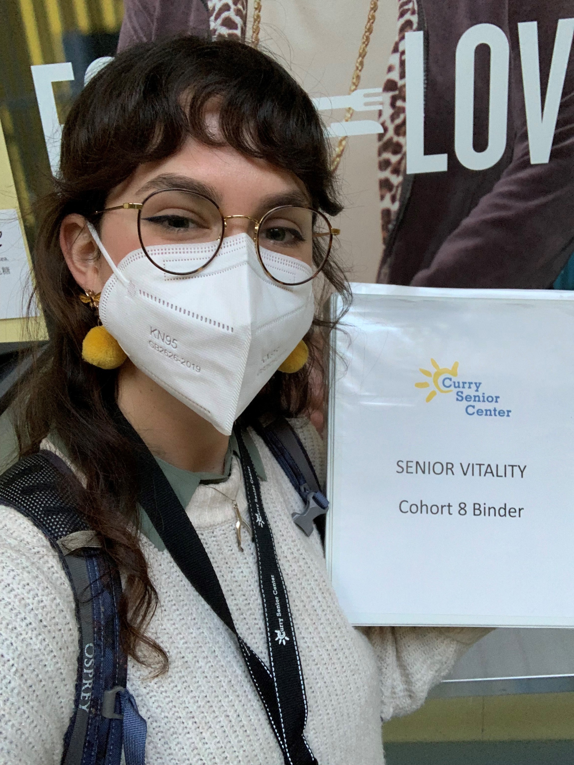 Sarah is facing the camera. She is holding a binder that reads "Curry Senior Center; Senior Vitality; Cohort 8 Binder". 