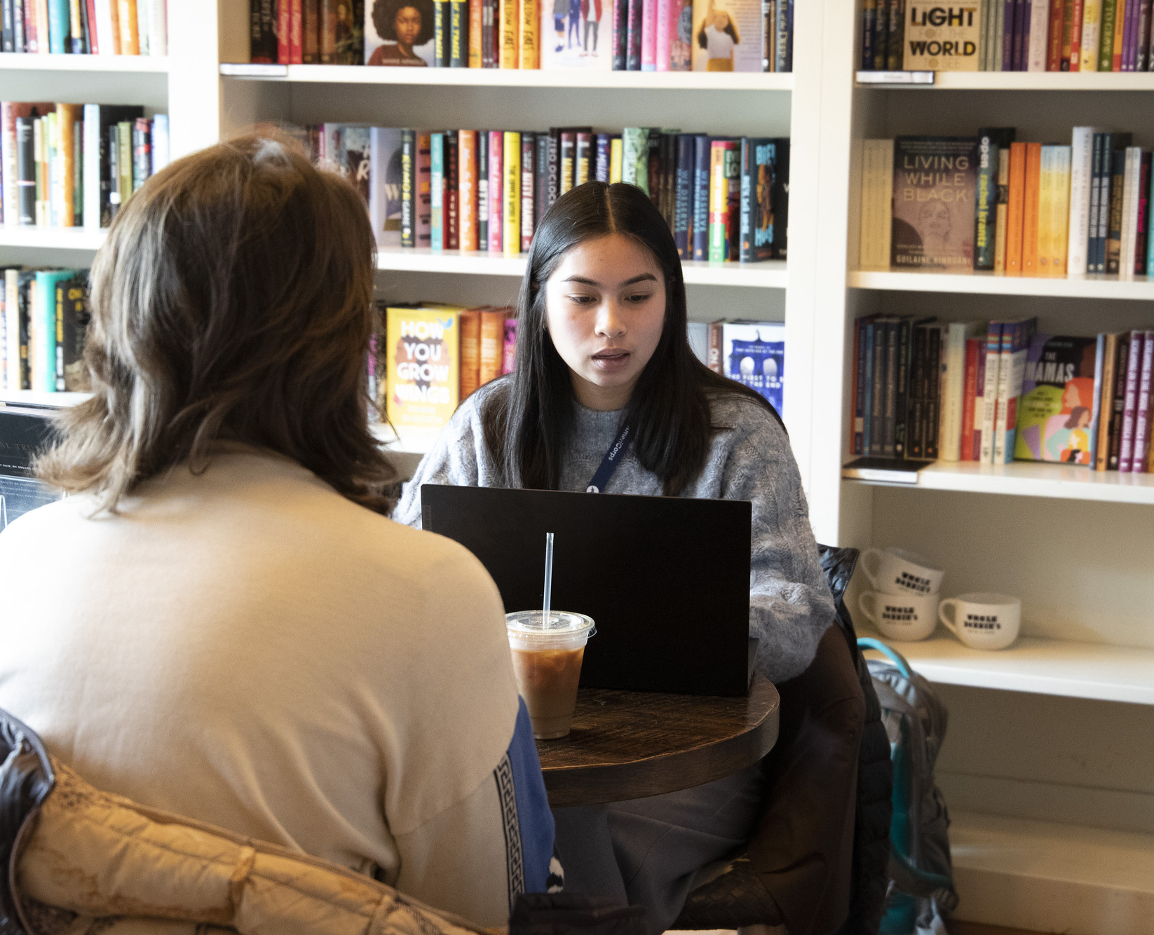 Patricia sitting at a table with her laptop in front of a bookshelf and there is a person facing her whose face we cannot see